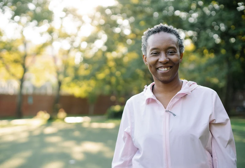 A smiling woman in a pink jacket stands in a sunny park