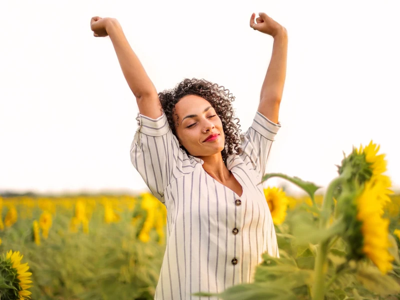 A smiling woman stands in a sunny field of sunflowers with her arms raised and her eyes closed