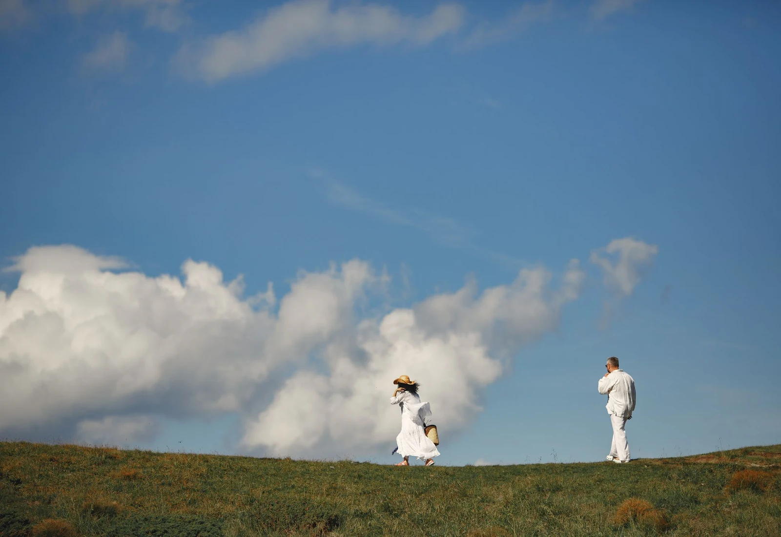 A man and a woman dressed in white walk along a sunny green hilltop under a bright blue sky with white clouds