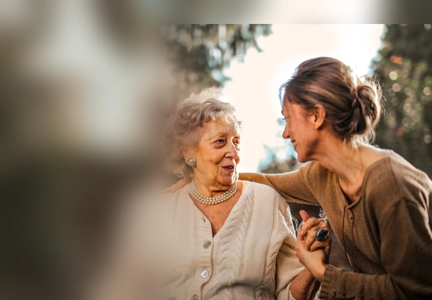 A smiling adult daughter embraces her happy older mother outdoors