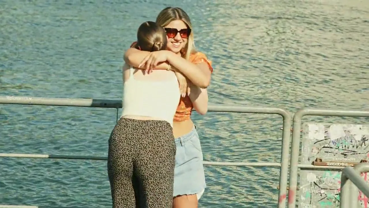 Two friends hug warmly on a sunny lakeside pier, then smile and hold hands